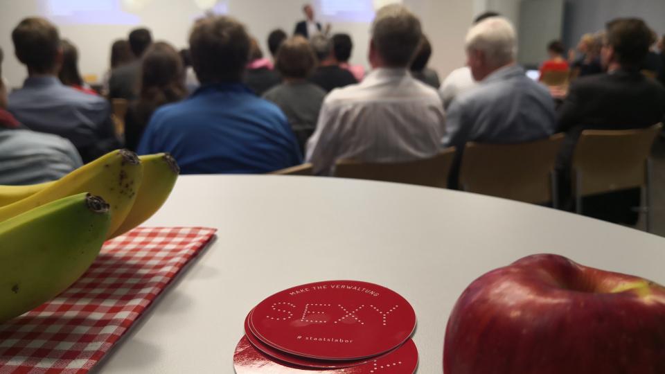 staatslabor Sticker und Obst mit Präsentation im Hintergrund